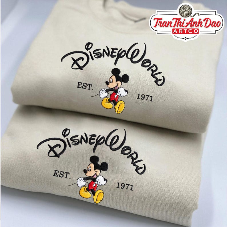 Puede incluir: Dos sudaderas beige con el logo de Disney World, un gr&aacute;fico de Mickey Mouse y el texto "EST. 1971". Las sudaderas est&aacute;n dobladas y apiladas. En la esquina superior derecha hay un logotipo con texto.