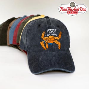 Puede incluir: Una fila de gorras de béisbol en varios colores, incluyendo negro, marrón y azul. La gorra delantera es gris oscuro con el texto "FIST MY BUMP!" y un gráfico de cangrejo de dibujos animados en naranja y azul.