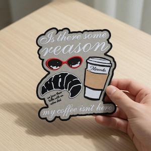 May include: Embroidered patch with a grey background and black border. The patch features a croissant, sunglasses, and a coffee cup with the name "Miranda." Text includes "Is there some reason," "my coffee isn't here," and "we four than talke with an extra shot."