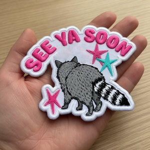Puede incluir: Parche bordado con un mapache gris con cola a rayas blancas y negras. El parche tiene las palabras "SEE YA SOON" en rosa, con estrellas rosas y una estrella turquesa, delineado en blanco.