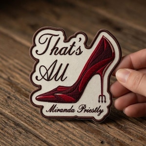 May include: Embroidered patch with a burgundy high-heeled shoe design. The patch has the text "That's All" and "Miranda Priestly" in a decorative font, outlined in brown. The patch is on a white background with a brown border.