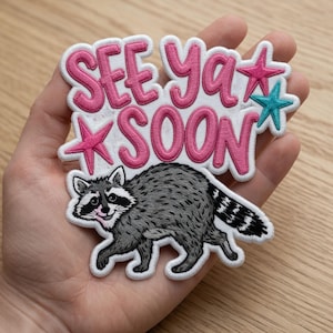 Puede incluir: Parche bordado con un mapache y la frase "SEE YA SOON" en letras rosas. El parche incluye estrellas rosas y turquesas. El mapache es gris y blanco con una cola a rayas negras.