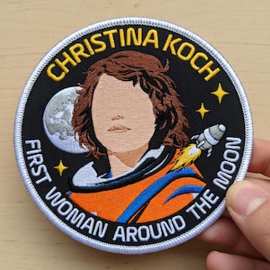 以下が含まれることがあります： 「CHRISTINA KOCH」と「FIRST WOMAN AROUND THE MOON」の文字が入った刺繍パッチ。肖像画、ロケット、月、星が黒い背景にデザインされています。