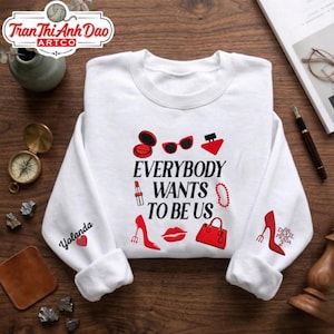 May include: White sweatshirt with the text "EVERYBODY WANTS TO BE US" in black, with red graphic icons of makeup, accessories, and high heels. The sleeve has the name "Yolanda" and a heart.