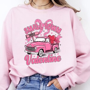 May include: Pink sweatshirt featuring a pink vintage truck carrying hearts. The graphic includes the words "Small Town Valentine" and a sign with the number "38863". The design is set against a light pink background.