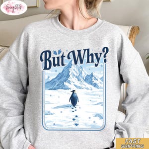 Puede incluir: Sudadera gris claro con un diseño de un pingüino caminando en la nieve hacia una cordillera. El texto "But Why?" está encima del diseño en azul. La sudadera es de estilo casual.