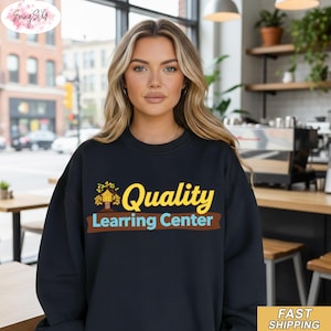 Könnte beinhalten: Ein schwarzes Sweatshirt mit dem Schriftzug "Quality Learning Center" in Gelb, Braun und Blau. Das Design beinhaltet eine Baum- und Hausgrafik. Das Sweatshirt wird von einer Person mit blonden Haaren getragen.