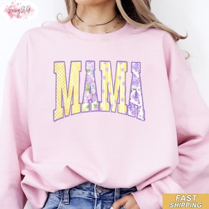 Może przedstawiać: Jasnoróżowa bluza z napisem "MAMA" w dużych literach z przodu. Litery są żółte w kropki, paski i kwiatowe wzory w kolorze fioletowym i białym. Swobodny styl.