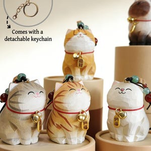 May include: Wooden cat figurines in various colors, including orange, gray, and white, each with a smiling face and a small bell. The figurines have a detachable keychain and are decorated with beads and a red cord.