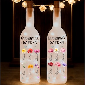 Op de afbeelding: Twee doorzichtige glazen flessen met kurken en witte lampjes erin. Elke fles heeft een bloemenmotief met de tekst "Grandma's Garden" en namen van familieleden gedrukt op de voorkant.