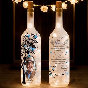 Puede incluir: Una botella de vidrio blanco con un tapón de corcho y una foto de un hombre con un sombrero. La botella está decorada con un árbol negro y mariposas azules. La botella tiene un mensaje que dice: "Él está en cada latido de mi corazón. El ángel de arriba, a quien extraño en cada momento de mi vida. El que quiero abrazar con fuerza. El que nunca está más lejos que un pensamiento, amado y recordado cada día. Él es mi esposo y mi ángel."
