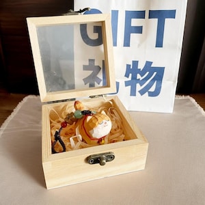 May include: A wooden box with a glass lid, containing a small wooden cat figurine with a beaded necklace. The box is sitting on a light brown surface. The box has a brass latch.