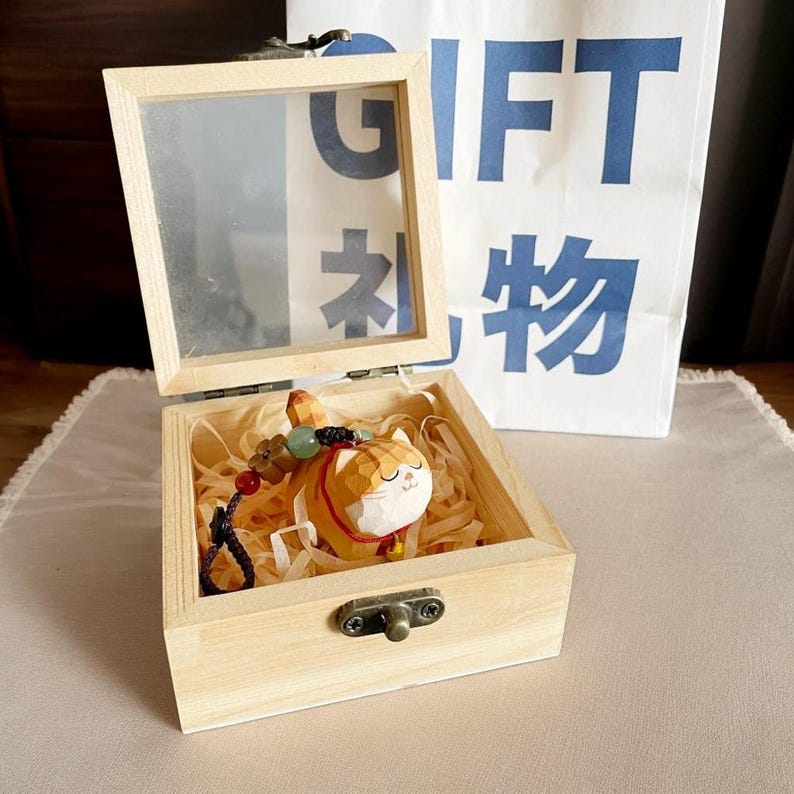 May include: A wooden box with a glass lid, containing a small wooden cat figurine with a beaded necklace. The box is sitting on a light brown surface. The box has a brass latch.