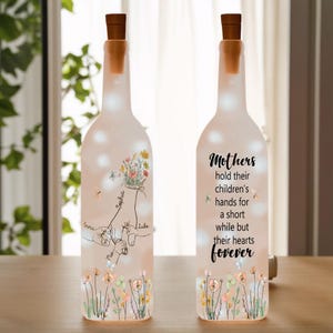 Op de afbeelding: Twee doorzichtige glazen flessen met kurkstoppen en witte lampjes erin. De flessen zijn versierd met een bloemenmotief en een citaat dat luidt: "Moeders houden de handen van hun kinderen vast voor een korte tijd, maar hun harten voor altijd". Eén fles heeft een handgetekende afbeelding van handen die elkaar vasthouden met de namen "Emma", "Sophia" en "Luke" die op de vingers zijn geschreven.