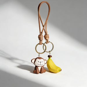 May include: A brown leather keychain with two brass rings. One ring holds a carved wooden monkey, the other a yellow banana. The monkey is brown and white, and the banana is yellow with a brown stem.