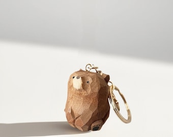Peut inclure: Un porte-clés en forme d'ours en bois avec un anneau doré. L'ours est sculpté dans du bois brun clair, avec une surface texturée et détaillée. Le visage de l'ours a une couleur de bois plus claire pour le museau et de petits yeux noirs.