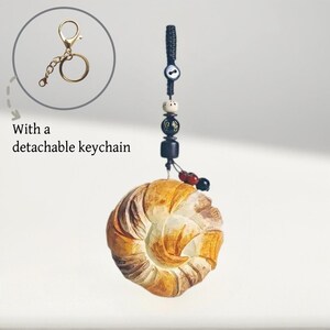 May include: A handcrafted keychain featuring a detailed, spiral-shaped pendant with brown and cream tones. The keychain includes a detachable gold-colored clasp and decorative beads. The text "With a detachable keychain" is also visible.