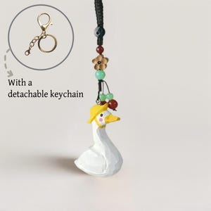 May include: A white goose ornament with a yellow hat, suspended from a black cord with decorative beads and a detachable gold keychain. The goose has a yellow beak and pink cheeks. Text in the image reads: "With a detachable keychain."