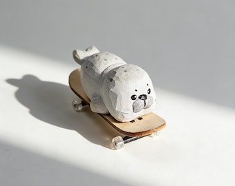 May include: A hand-carved wooden seal figurine with a light gray finish and dark gray spots, featuring a painted face with black eyes and a gray snout. The seal is positioned on top of a small, light-colored wooden toy skateboard with wheels.