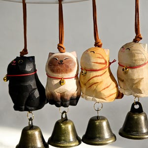 May include: Four wooden cat ornaments with bronze bells hanging from brown leather cords. The cats are painted black, brown, orange, and beige, each with a red collar and a small gold bell. The ornaments are suspended from a white bar.