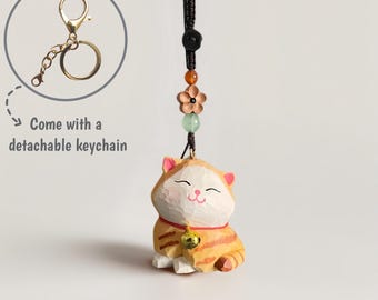 May include: A wooden cat figurine with orange and white stripes, a red collar, and a gold bell. The cat charm has a detachable gold keychain and is attached to a black cord with decorative beads and a flower. The text in the image says "Come with a detachable keychain."