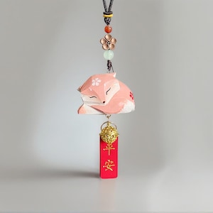 Könnte beinhalten: Hängeornament mit einem schlafenden rosa Fuchs mit floralen Akzenten. Der Fuchs hängt an einer schwarzen Schnur mit dekorativen Perlen und einer Blume. Ein rotes Etikett mit goldenen chinesischen Schriftzeichen hängt darunter.