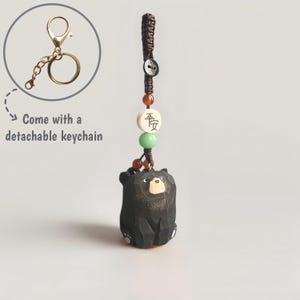 May include: A black carved bear keychain with a detachable gold-colored keychain. The bear has a light-colored face and feet. The keychain includes a brown cord, beads, and a button. The text "Come with a detachable keychain" is also visible.