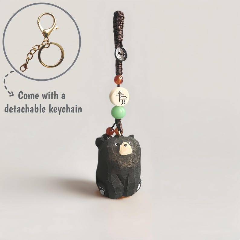 May include: A black carved bear keychain with a detachable gold-colored keychain. The bear has a light-colored face and feet. The keychain includes a brown cord, beads, and a button. The text "Come with a detachable keychain" is also visible.