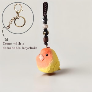 May include: A hand-carved wooden bird keychain in shades of yellow and orange, with a detachable gold-colored keychain. The bird has a detailed, textured appearance and is suspended from a beaded cord. The text "Come with a detachable keychain" is also visible.