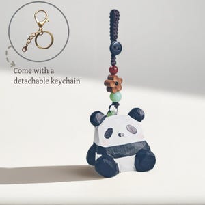 May include: A panda bear keychain with a detachable gold-colored keychain. The panda is black and white with a small pink blush on its cheeks. The keychain has a decorative cord with beads and a wooden flower.