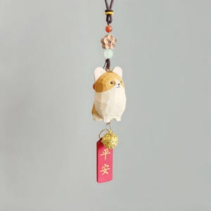 Könnte beinhalten: Ein Holzanhänger in Form eines Corgi-Hundes mit braun-weißem Farbschema, der an einer braunen Schnur hängt. Der Anhänger ist mit einer Blume, Perlen und einem roten Anhänger mit chinesischen Schriftzeichen verziert. Unterhalb des Anhängers hängt eine goldfarbene Glocke.