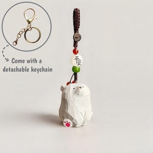 May include: A white and gray carved bear keychain with a detachable gold-colored keychain. The bear has pink paw pads and is attached to a brown cord with red, white, and green beads. The white bead has black Chinese characters.