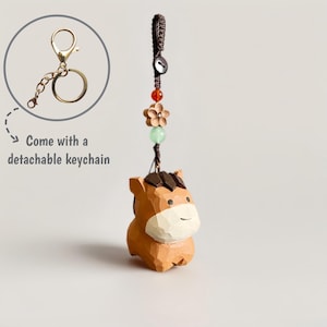 May include: A wooden horse keychain with a detachable gold-colored keychain. The horse is light brown with a dark brown mane and a smiling face. The keychain includes a decorative cord with beads and a flower.