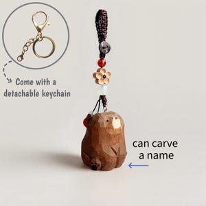 May include: A brown, carved wooden bear keychain with a detachable gold-colored keychain. The bear has a detailed, textured surface and is adorned with decorative beads and a flower. Text on the image says "can carve a name".