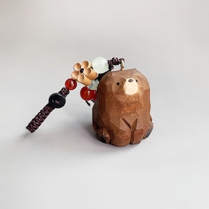 May include: A carved wooden bear keychain with a dark brown braided cord and decorative beads. The bear is a light brown color with a carved design. The keychain also features a wooden flower and red and white beads.