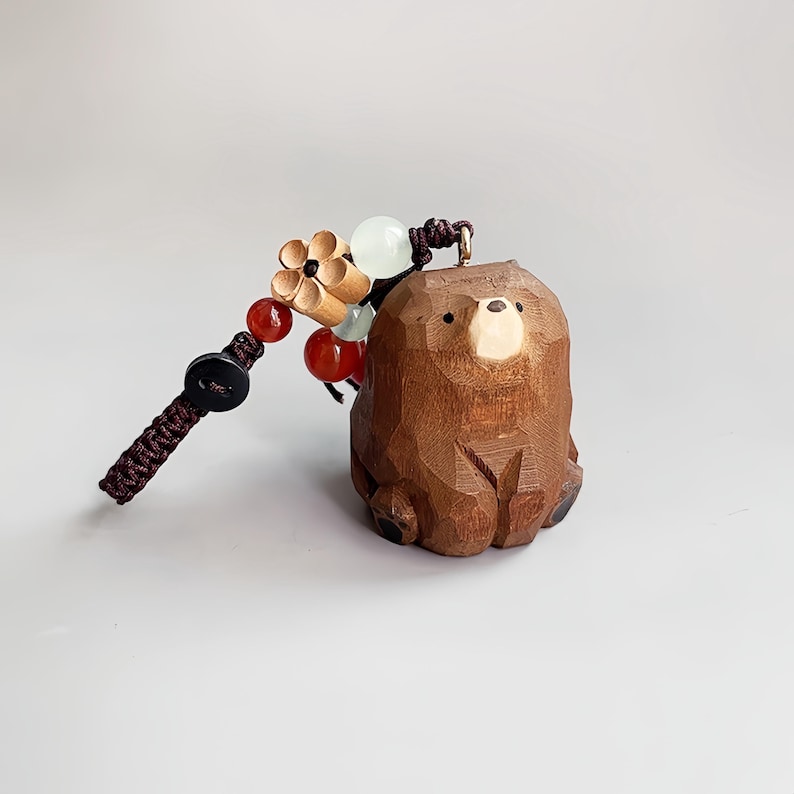 May include: A carved wooden bear keychain with a dark brown braided cord and decorative beads. The bear is a light brown color with a carved design. The keychain also features a wooden flower and red and white beads.