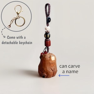 May include: A wooden bear keychain with a detachable gold-colored keychain. The bear is carved from wood and has a dark brown finish. The keychain includes decorative beads and the text "can carve a name".
