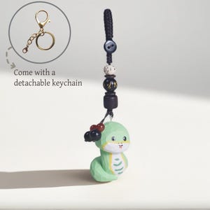 May include: A green snake-shaped keyring with a detachable gold-coloured keyring. The snake has a light green body with yellow accents and black and red beads. The keyring has a black woven cord with decorative beads.