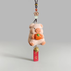 May include: A pink wooden pig figurine holding an orange fruit, hanging from a brown cord with green and brown beads. The pig has a gold bell and a red tag with gold Chinese characters that read "Peace" hanging from it.