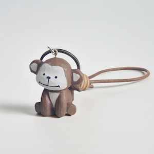 Könnte beinhalten: Ein brauner Holz-Affen-Schlüsselanhänger mit weißem Gesicht und Ohren. Der Affe sitzt mit seinen Armen an den Seiten. Der Schlüsselanhänger hat einen Metallring und einen braunen Lederriemen. Der Affe ist eine geschnitzte Figur.