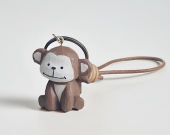 May include: A brown wooden monkey keychain with a white face and ears. The monkey is sitting with its arms at its sides. The keychain has a metal ring and a brown leather strap. The monkey is a carved figurine.