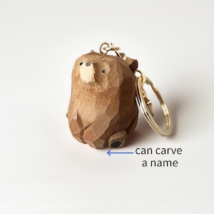 May include: A wooden bear keychain with a gold-colored keyring. The bear is carved from wood and has a light-colored snout and small black eyes. The text "can carve a name" is below the bear.