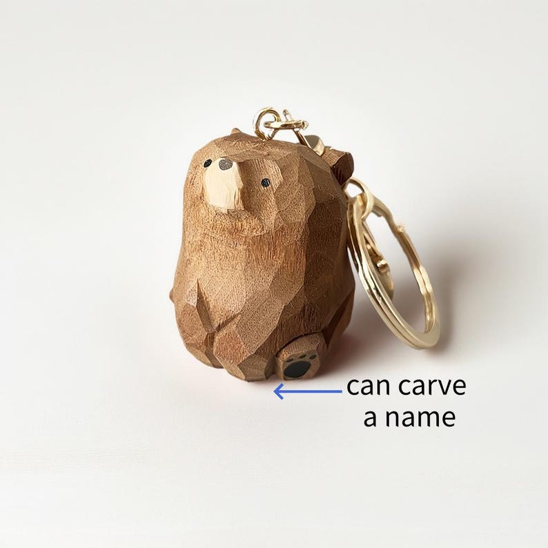 May include: A wooden bear keychain with a gold-colored keyring. The bear is carved from wood and has a light-colored snout and small black eyes. The text "can carve a name" is below the bear.