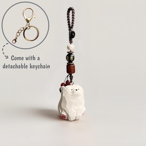 May include: A white bear keychain with a detachable gold-colored keychain. The bear has a carved appearance with pink accents on its feet. The keychain includes a beaded strand with wooden and decorative beads. The text "Come with a detachable keychain" is also visible.