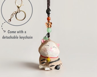 May include: A wooden cat figurine with a detachable gold-coloured keychain. The cat is tan with black stripes and a red collar with a small gold bell. The keychain has a decorative beaded strand with a flower and green beads. Text reads: "Come with a detachable keychain."