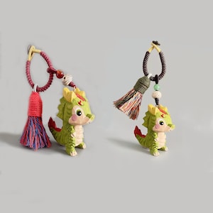 Op de afbeelding: Twee draakvormige sleutelhangers. De draken zijn groen met gele accenten en rode staarten. Elke sleutelhanger heeft een kwast en decoratieve kralen. Eén kwast is rood en blauw, de andere is veelkleurig.