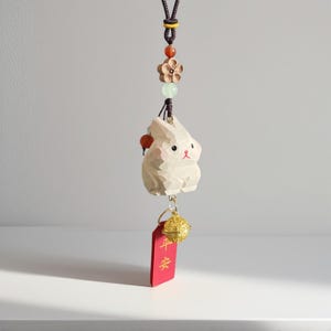 Puede incluir: Un amuleto de conejo de madera tallada con una etiqueta roja y una campana dorada. El conejo es blanco con ojos negros y mejillas rosadas. La etiqueta roja tiene caracteres chinos dorados. Cuelga de un cordón marrón.