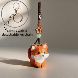 May include: A wooden fox keyring with a detachable gold-coloured keyring. The fox is orange and white with a detailed carved design. The keyring includes decorative beads and a wooden flower. The text says "Comes with a detachable keyring."