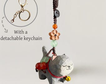 May include: A grey cat-shaped keyring with a detachable gold-coloured keyring. The cat has a red collar with a small gold bell. The keyring is decorated with beads and a wooden flower.