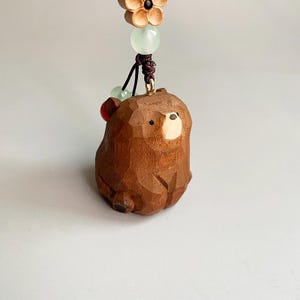 May include: A carved wooden bear figurine with a light brown finish. The bear is suspended by a dark brown cord, adorned with green and red beads, and a wooden flower. The bear's face has a lighter wood color.